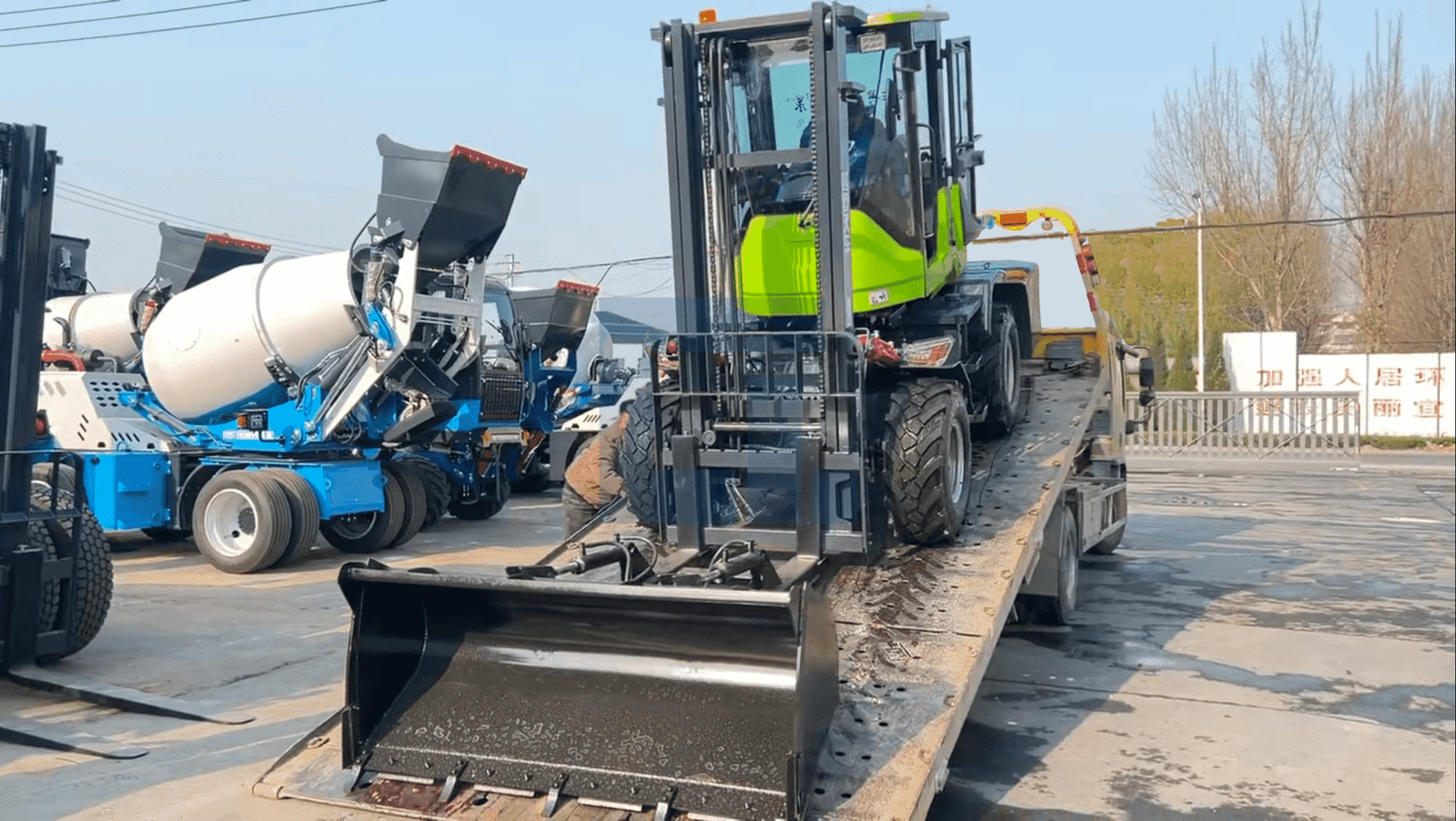 HIGHLAND HEAVY MACHINERY green rough terrain forklift being loaded onto a red flatbed trailer in a wet factory yard, with blue self loading concrete mixer trucks in the background and a worker guiding the process.