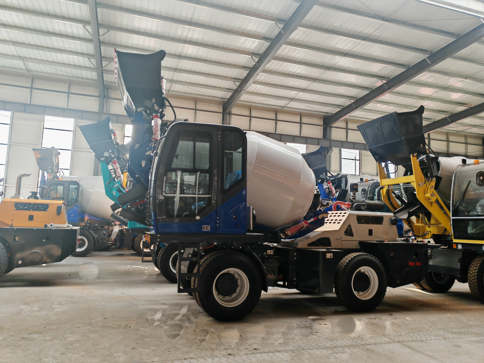 HIGHLAND HEAVY MACHINERY indoor factory workshop with multiple heavy equipment units, including a blue and white self loading concrete mixer truck in the foreground and a yellow wheel loader in the background, ready for assembly and inspection.