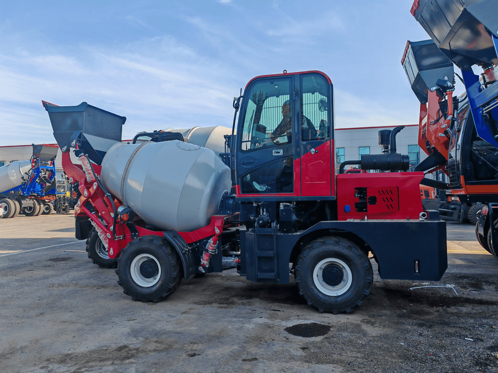 HIGHLAND HEAVY MACHINERY red self loading concrete mixer truck with white mixing drum, operator seated in the enclosed cabin, left side rear three-quarter view on a wet concrete service yard with other mixer trucks in the background.