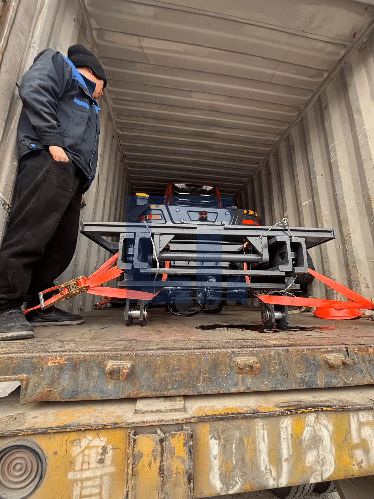 HIGHLAND HEAVY MACHINERY worker carefully securing a heavy-duty off-road forklift attachment with high-tensile orange ratchet straps on the container floor for safe international shipping.