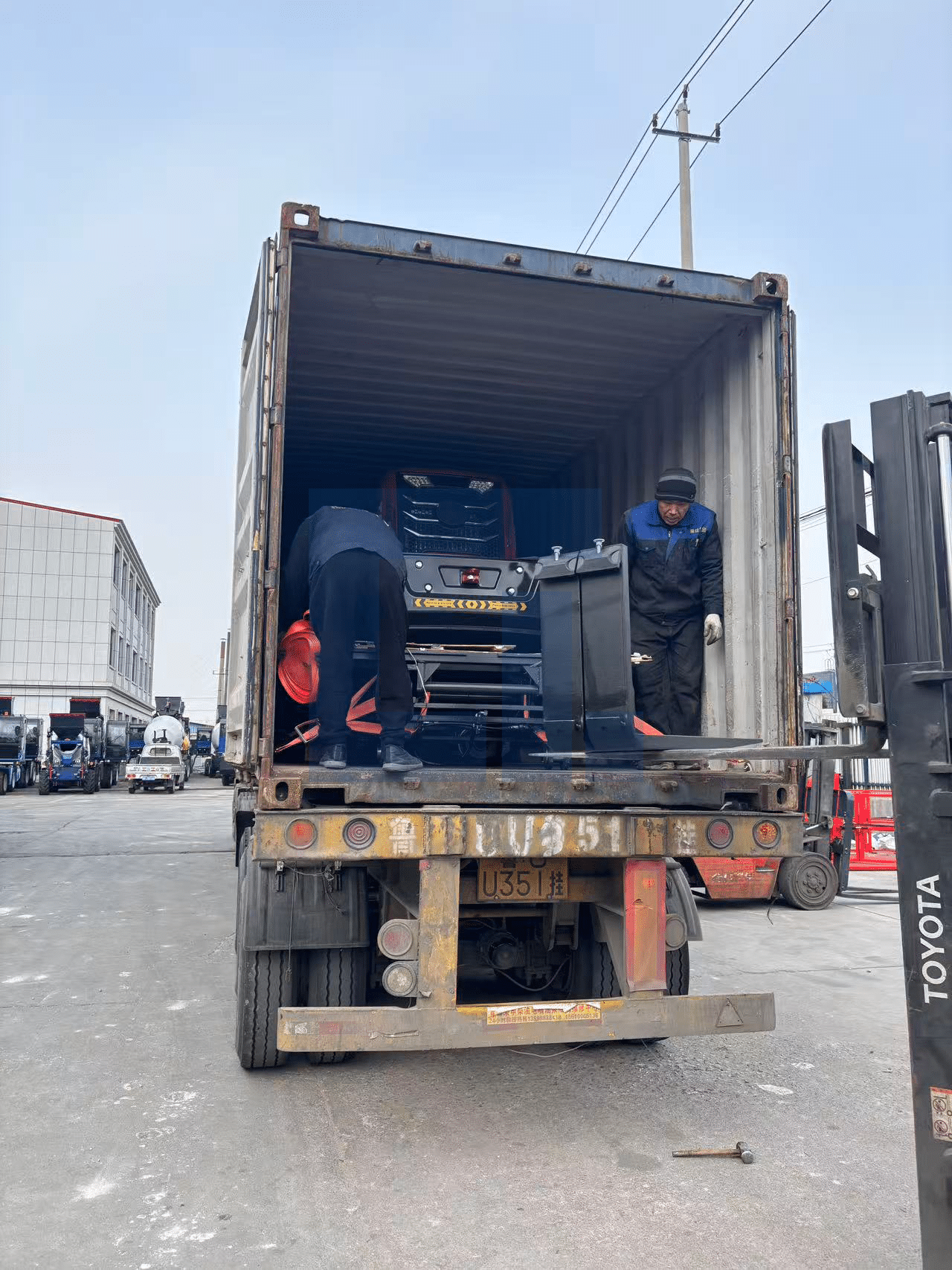 HIGHLAND HEAVY MACHINERY workers carefully securing a heavy-duty black off-road forklift attachment inside a shipping container, optimizing space and ensuring safe transport for global export.