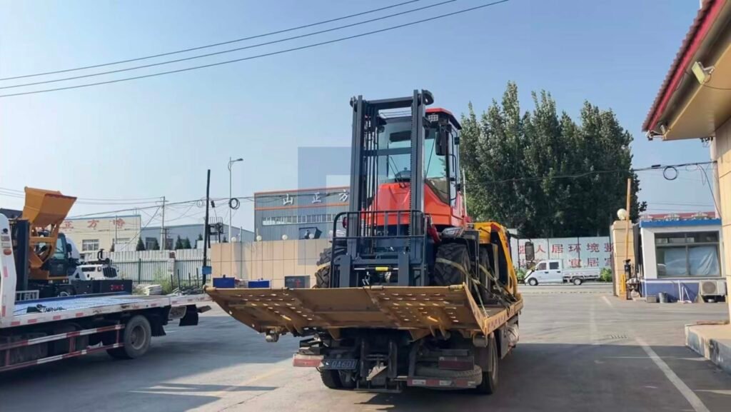 HIGHLAND HEAVY MACHINERY orange rough terrain forklift with a load on its forks, being carefully loaded onto a flatbed trailer at the factory dispatch yard for global shipping.