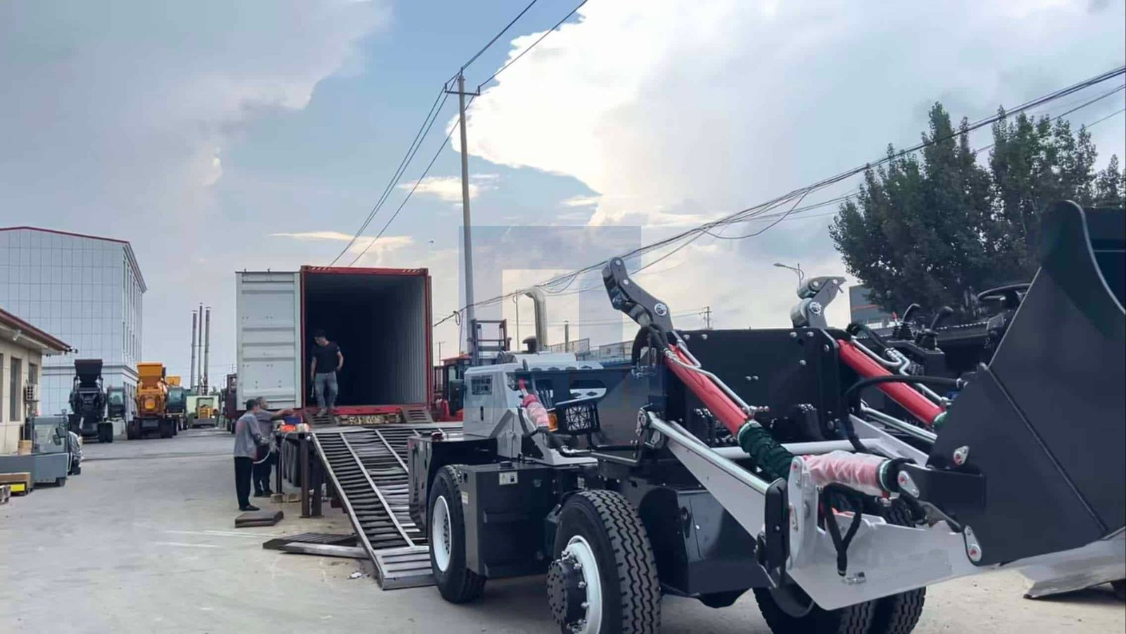 HIGHLAND HEAVY MACHINERY self loading concrete mixer truck with raised red hydraulic cylinders and large bucket, positioned near a shipping container with a metal loading ramp in a wet factory yard, preparing for export dispatch.
