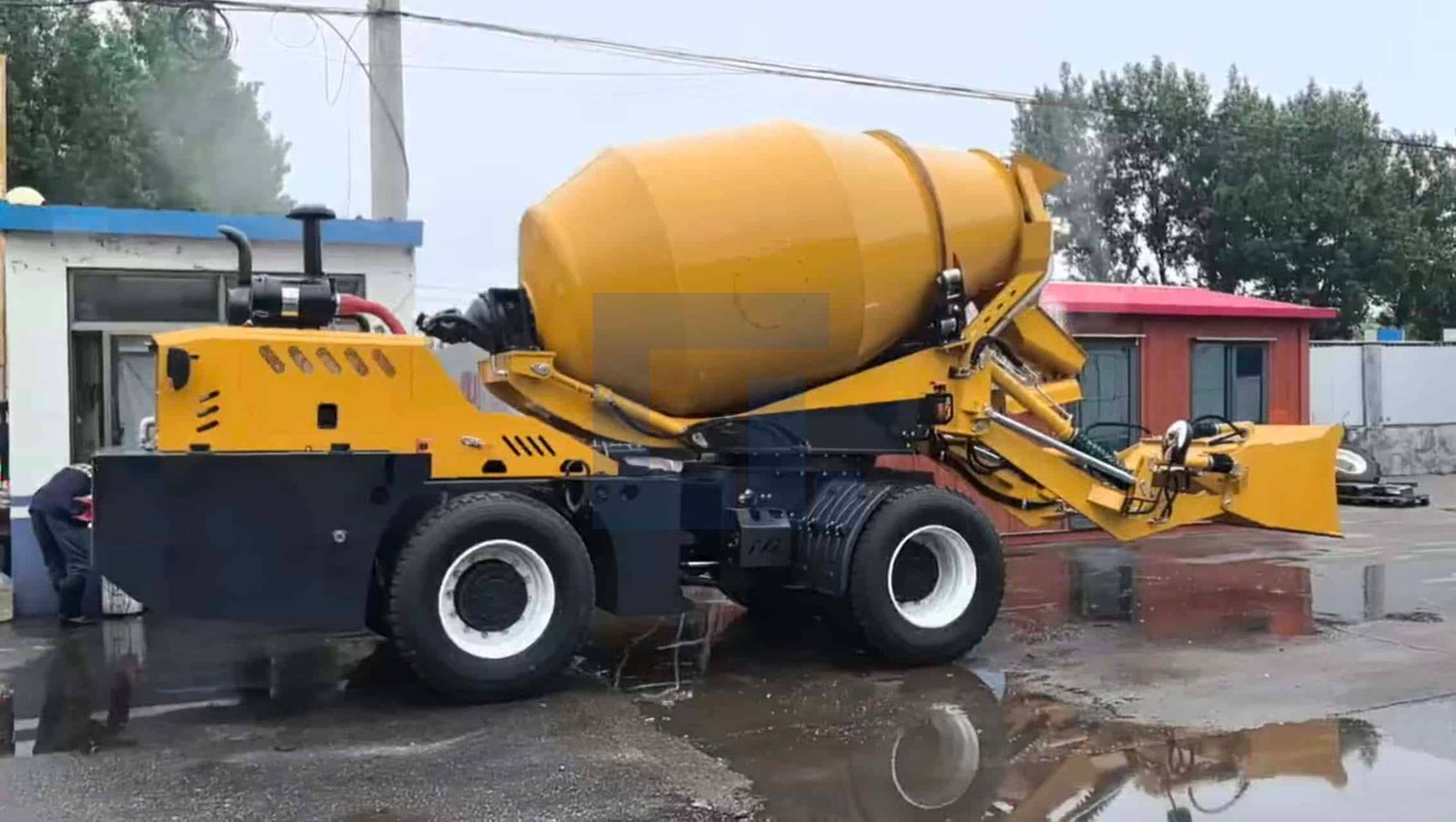 HIGHLAND HEAVY MACHINERY yellow self loading concrete mixer truck side view with front loader bucket slightly raised, parked on a wet reflective yard, ready for material scooping and on-site concrete batching.